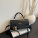 A black leather handbag with a top handle and gold-tone hardware, styled on a white book next to a white textured vase with dried branches against a light wall.