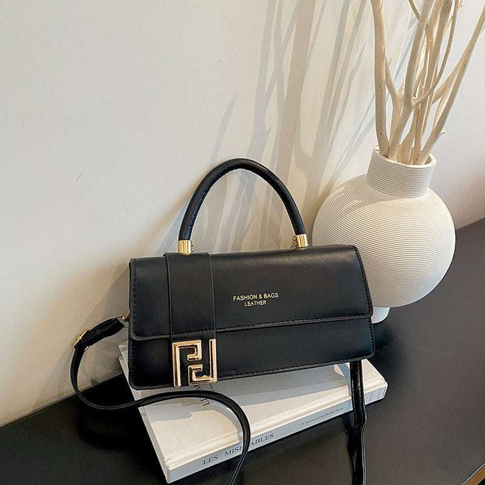 A black leather handbag with a top handle and gold-tone hardware, styled on a white book next to a white textured vase with dried branches against a light wall.