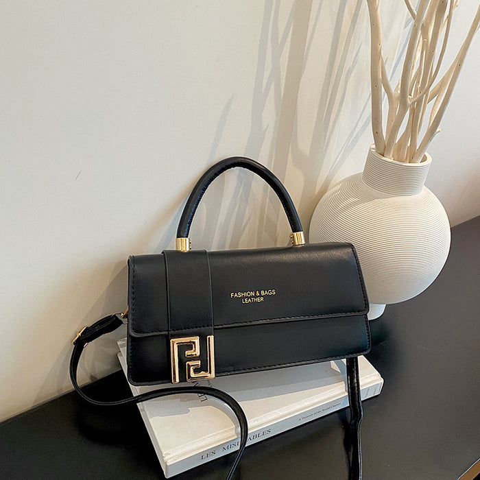 A black leather handbag with a top handle and gold-tone hardware, styled on a white book next to a white textured vase with dried branches against a light wall.