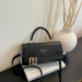 A black leather handbag with a top handle and gold-tone hardware, styled on a white book next to a white textured vase with dried branches against a light wall.