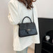 A woman wearing a white blazer carries a black crocodile-embossed top-handle handbag with a shoulder strap and gold-tone hardware.