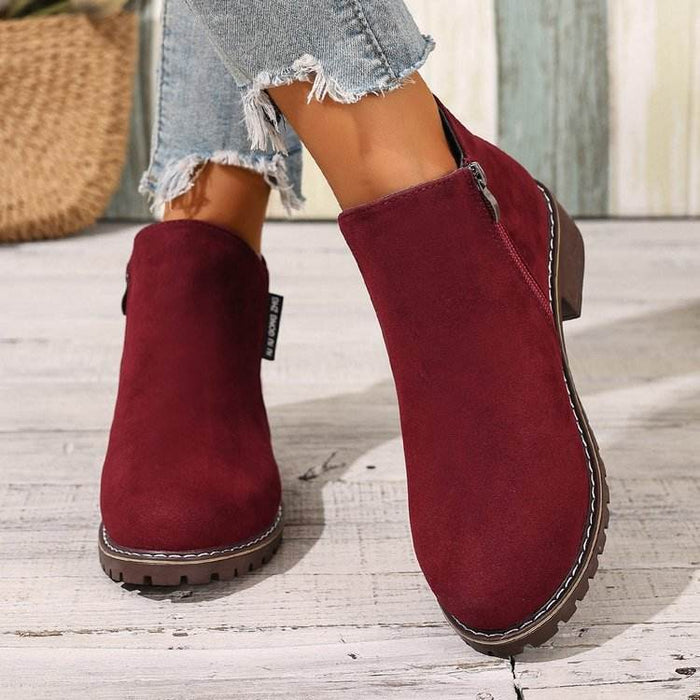 Red suede ankle boots worn with denim jeans on a wooden floor.
