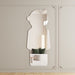 A vertically oriented wall mirror with an irregular silhouette shaped like a person, mounted on a light beige paneled wall. The mirror reflects a softly lit interior with a white sofa, dark green pillows, and a decorative lamp.