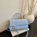 A light blue faux leather handbag with a top handle, a matching shoulder strap, and a gold-tone geometric frame clasp, styled on a white book next to a white vase with dried branches.