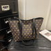 A brown canvas tote bag with a repeating gold monogram pattern and black leather trim, resting on a dark wooden surface next to a black cabinet and a white book.