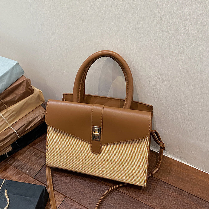 A structured handbag with a tan leather top handle and flap, combined with a beige woven body, resting on a wooden floor against a plain wall.