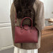 A woman wearing a brown tweed blazer carries a burgundy pebbled leather handbag with top handles and a shoulder strap.