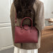 A woman wearing a brown tweed blazer carries a burgundy pebbled leather handbag with top handles and a shoulder strap.