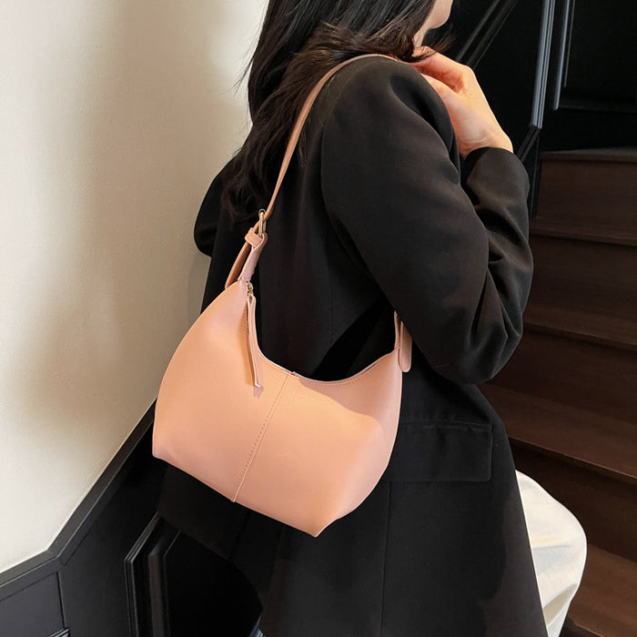 A woman wearing a black coat carries a soft blush pink crescent-shaped shoulder bag with a matching strap in an indoor setting.
