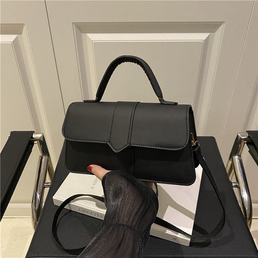 A black structured handbag with a top handle and shoulder strap, displayed on a chair in an indoor setting. A hand with red nail polish rests on the bag, which is propped against a white book.