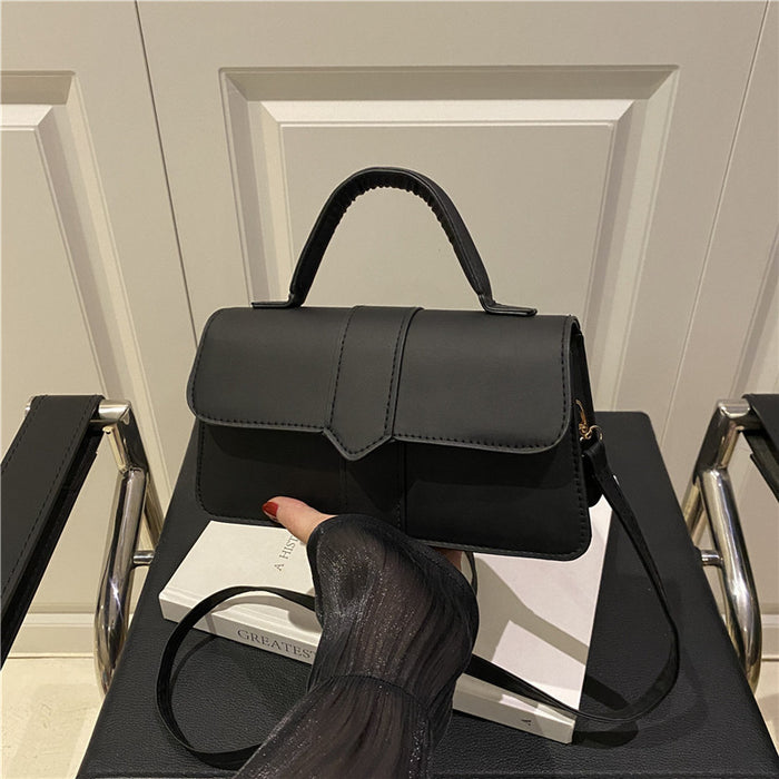 A black structured handbag with a top handle and shoulder strap, displayed on a chair in an indoor setting. A hand with red nail polish rests on the bag, which is propped against a white book.