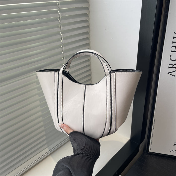 A glossy white leather handbag with black piping and double top handles, held by a hand wearing a black long-sleeve top in front of window blinds.