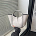A glossy white leather handbag with black piping and double top handles, held by a hand wearing a black long-sleeve top in front of window blinds.