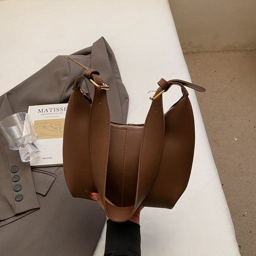 A brown leather shoulder bag with gold-tone hardware, held by a hand over a gray blazer and a book on a white surface.