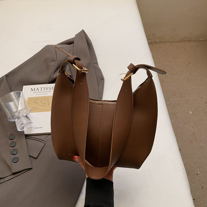 A brown leather shoulder bag with gold-tone hardware, held by a hand over a gray blazer and a book on a white surface.
