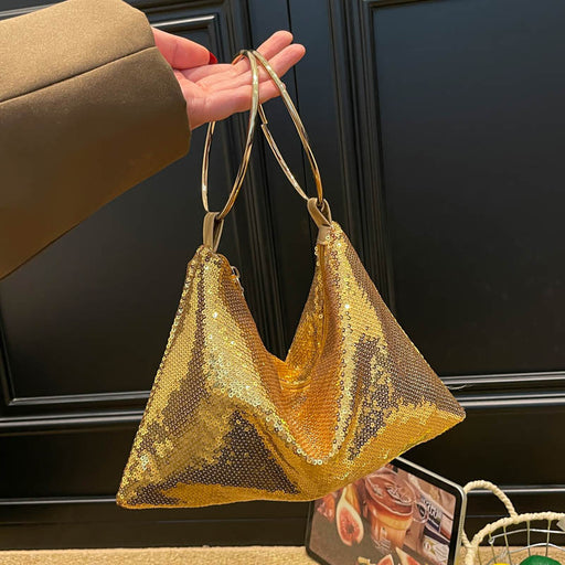 A gold sequined slouchy shoulder bag with double metallic gold handles, held by a hand wearing a brown sleeve against a dark paneled background.