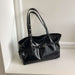 A black patent leather tote bag with double shoulder straps and a front strap detail, resting on a white shelf against a white wall.