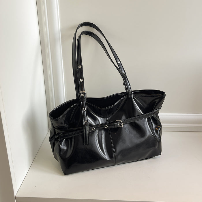 A black patent leather tote bag with double shoulder straps and a front strap detail, resting on a white shelf against a white wall.