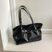A black patent leather tote bag with double shoulder straps and a front strap detail, resting on a white shelf against a white wall.