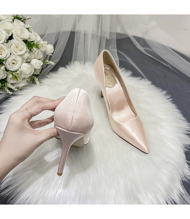 Pink high-heeled shoes being held by a hand on a white fur surface with a bouquet of white roses and a veil in the background.