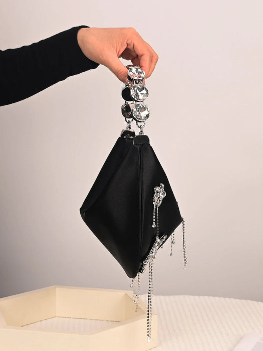 Black handbag with decorative chain and gemstones held by a hand against a neutral background
