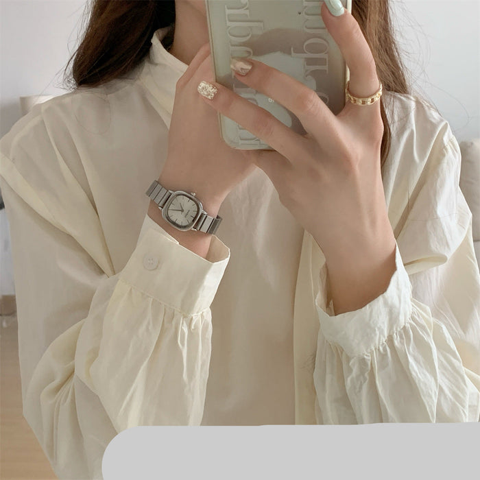 A woman takes a mirror selfie wearing a cream-colored long-sleeve blouse with gathered cuffs, a silver-toned metal watch on her wrist, and gold rings on her fingers.