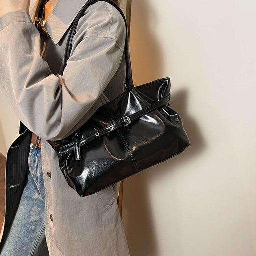 A person wearing a light gray oversized shirt and blue jeans carries a shiny black shoulder bag with a strap and front buckle detail against a plain indoor wall.