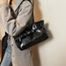A person wearing a light gray oversized shirt and blue jeans carries a shiny black shoulder bag with a strap and front buckle detail against a plain indoor wall.