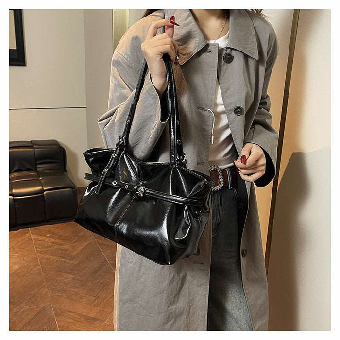 A person wearing a light gray trench coat and dark jeans carries a shiny black shoulder bag with a structured top handle and a wide strap detail, standing indoors near a mirror.