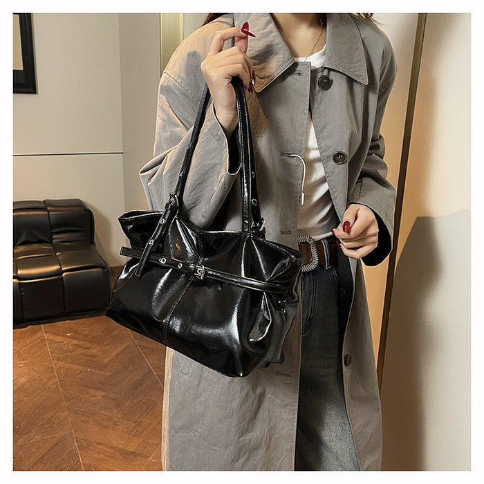 A person wearing a light gray trench coat and dark jeans carries a shiny black shoulder bag with a structured top handle and a wide strap detail, standing indoors near a mirror.