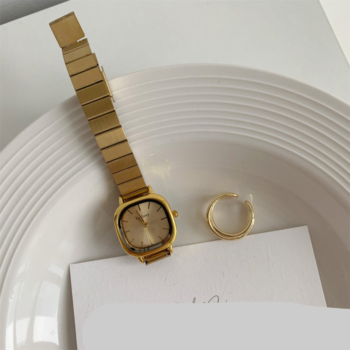 A gold-tone metal link watch with a square bezel and matching gold dial, displayed on a white plate alongside a matching gold ring and a card.