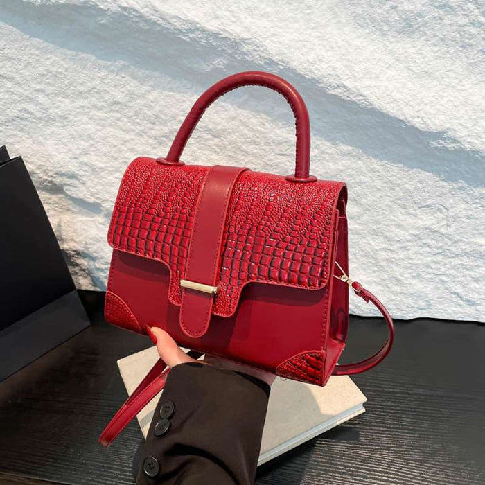 A bright red handbag with a crocodile-embossed texture on the front flap and smooth red leather trim, held by a hand over a stack of books against a textured white wall.