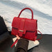 A bright red handbag with a crocodile-embossed texture on the front flap and smooth red leather trim, held by a hand over a stack of books against a textured white wall.