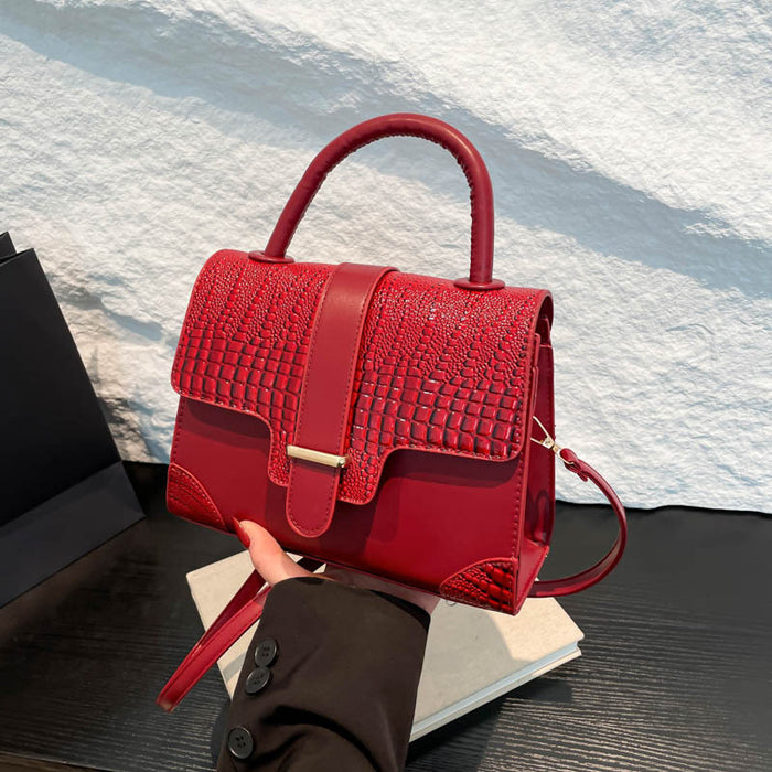 A bright red handbag with a crocodile-embossed texture on the front flap and smooth red leather trim, held by a hand over a stack of books against a textured white wall.