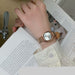 A person wearing a rose gold-toned metal watch with a square face and matching metal bracelet, resting their hand on an open book.