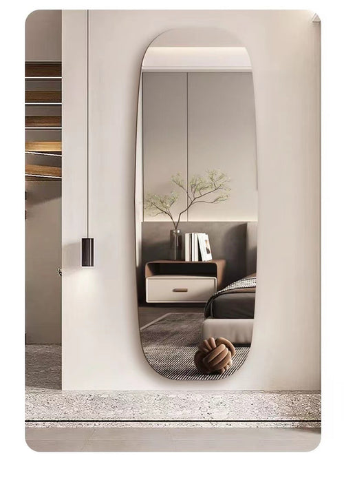 An arched, full-length wall mirror with a rounded top, mounted on a beige wall in a modern bedroom setting. The mirror reflects a neutral-toned bed, side table, and vase with branches.