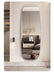 An arched, full-length wall mirror with a rounded top, mounted on a beige wall in a modern bedroom setting. The mirror reflects a neutral-toned bed, side table, and vase with branches.