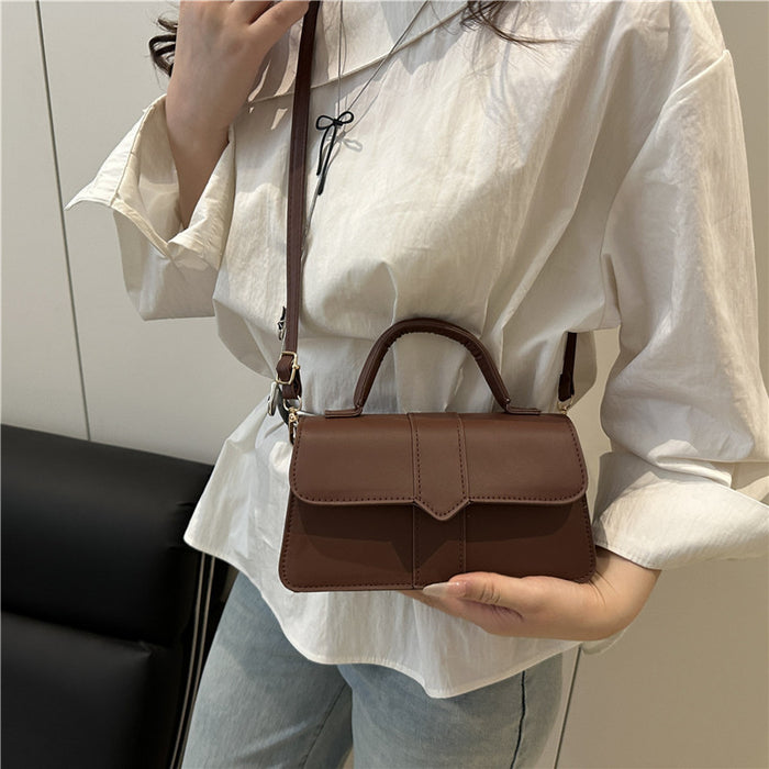 A person wearing a white blouse and light blue jeans carries a small brown top-handle crossbody bag with a matching shoulder strap. The bag has a structured rectangular shape, a top handle, a front flap, and a zipper closure.