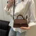 A person wearing a white blouse and light blue jeans carries a small brown top-handle crossbody bag with a matching shoulder strap. The bag has a structured rectangular shape, a top handle, a front flap, and a zipper closure.