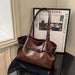 A brown leather shoulder bag with a structured slouchy shape, dual top handles, and a front strap detail, displayed on a black table next to a framed print.