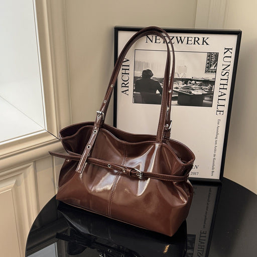 A brown leather shoulder bag with a structured slouchy shape, dual top handles, and a front strap detail, displayed on a black table next to a framed print.