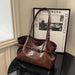 A brown leather shoulder bag with a structured slouchy shape, dual top handles, and a front strap detail, displayed on a black table next to a framed print.