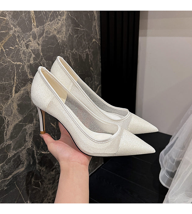 White high-heeled shoes held by a hand against a marble wall.