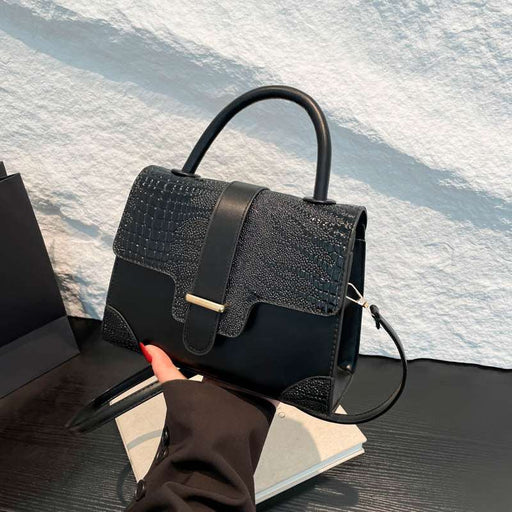 A structured black handbag with a crocodile-embossed flap and smooth leather trim, held by a hand over a white textured wall and dark tabletop.