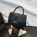 A structured black handbag with a crocodile-embossed flap and smooth leather trim, held by a hand over a white textured wall and dark tabletop.