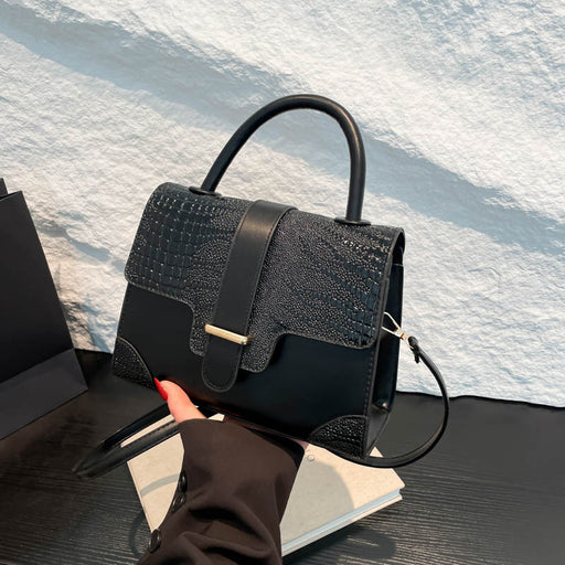 A structured black handbag with a crocodile-embossed flap and smooth leather trim, held by a hand over a white textured wall and dark tabletop.