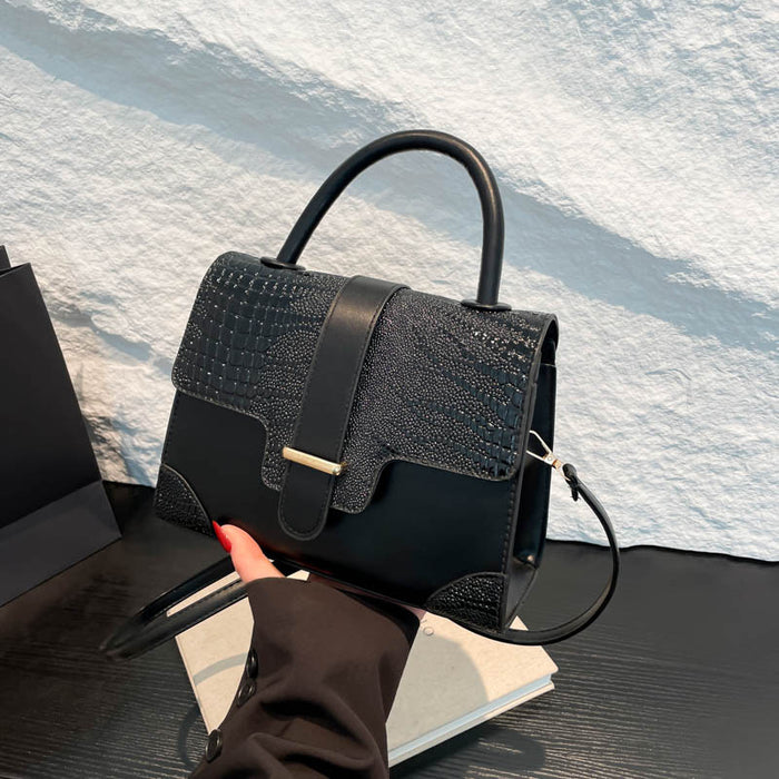 A structured black handbag with a crocodile-embossed flap and smooth leather trim, held by a hand over a white textured wall and dark tabletop.