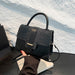 A structured black handbag with a crocodile-embossed flap and smooth leather trim, held by a hand over a white textured wall and dark tabletop.