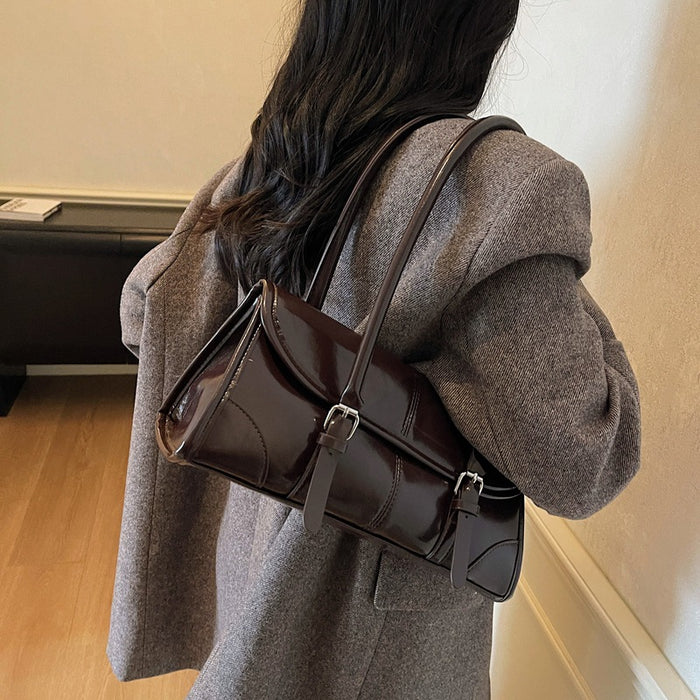 A person wearing a gray coat carries a dark brown glossy leather shoulder bag with silver buckle details, shown from the back in an indoor setting.