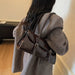 A person wearing a gray coat carries a dark brown glossy leather shoulder bag with silver buckle details, shown from the back in an indoor setting.